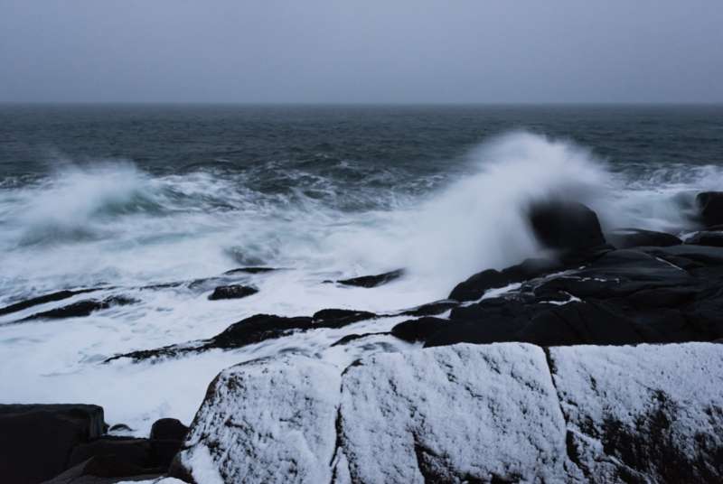 Heavy, dark gray storm clouds hang over dark, cold water during a winter storm in the north Atlantic. Heavy surf batters frost-covered rocks.